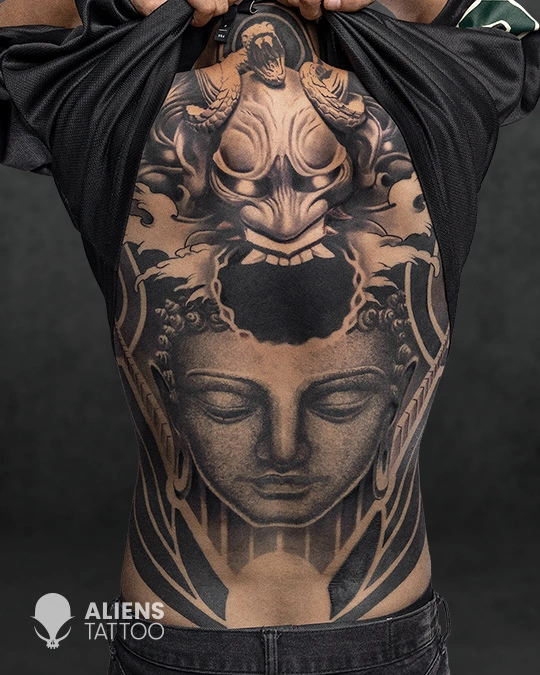 Black and grey full back tattoo featuring a Buddha face with a fierce demon mask above, detailed shading and symmetrical design