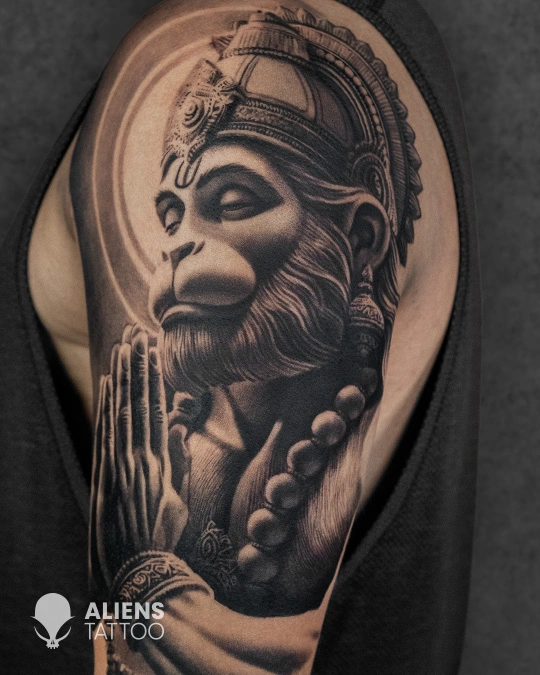 Hyper realistic black and grey Lord Hanuman tattoo in a prayer pose with detailed facial features and divine aura on upper arm, created by Aliens Tattoo, the best tattoo studio in pune.