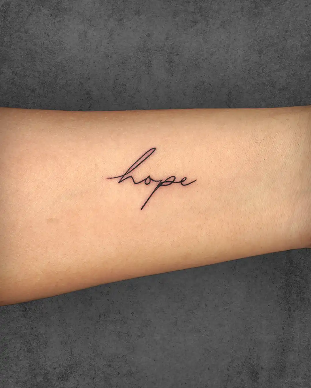 Simple "Hope" tattoo on a woman's inner forearm, representing optimism and resilience in a clean, cursive script.