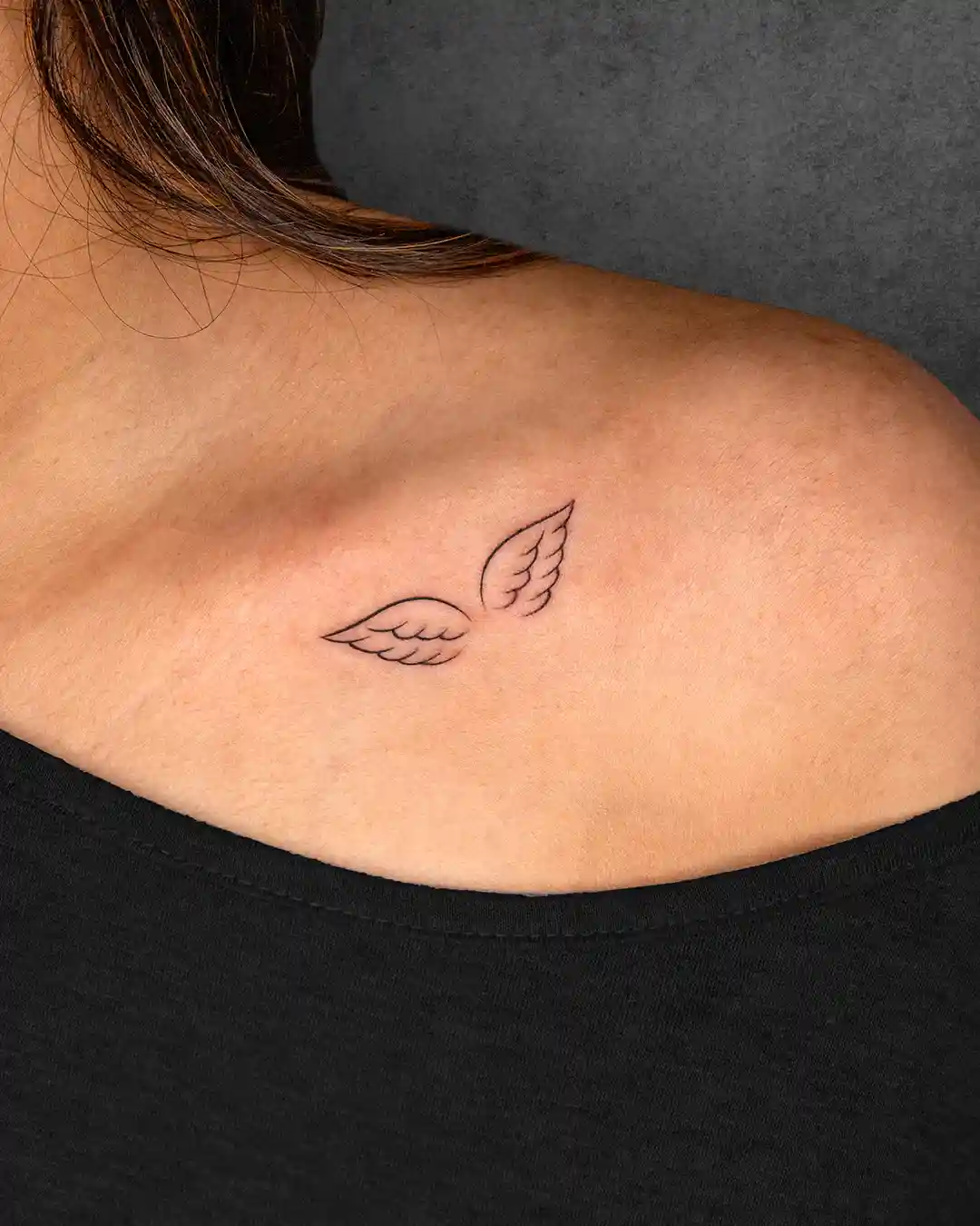 Small angel wings tattoo on a woman's shoulder blade, symbolizing freedom or protection with a minimalistic design.