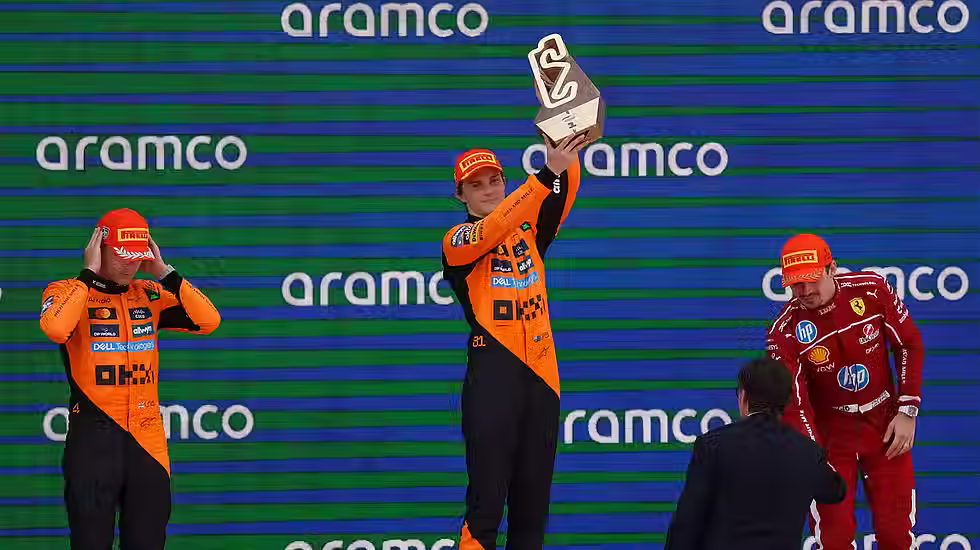A male race car driver in a papaya-colored racing suit holds up a first place trophy on the podium while his teammate stands to the left in second place and a red-suited driver stands on the right in third place.