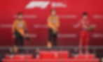 Three male racecar drivers stand on a bright red podium and backdrop with white writing.