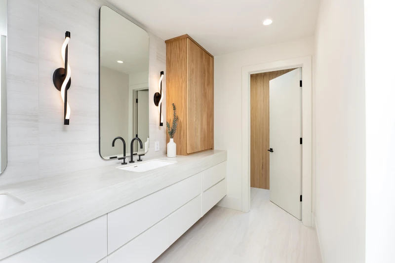 White bathroom vanity with a tall mirror, curved wall lights and a wood-grain cabinet.