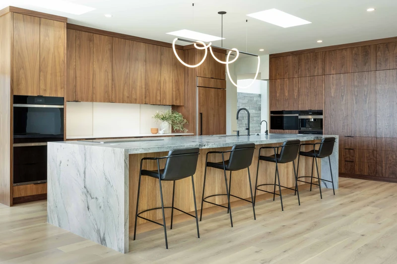 Veined waterfall island and wood-grain kitchen cabinets beneath a curved pendant light.