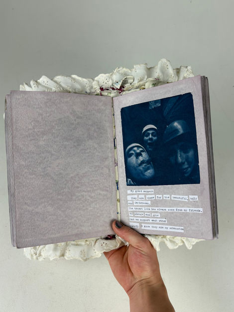 The image shows a hand holding a book open to a page in the middle. Around the pages you can see fabric ruffles. The pages themselves have a slight purple hue, and the visible page has a deep blue image with text underneath. The image shows three girls making faces into the camera. They crowd the camera, making the two girls in front look very large and the one in the back small. The background is too dark to see anything. The text is arranged so that it is aligned on the left side. It is made up of collaged type writer words. 
