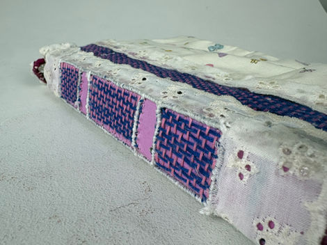 The image shows a closeup image of the book’s spine. There are three blocks of visible weaving on the spine showing the pink and blue stitching. On he boarders of the three blocks, there is white waxed linen that is creating a solid white boarder around the woven blocks. The very bottom edges and sides are covered in thin lace ruffles. 