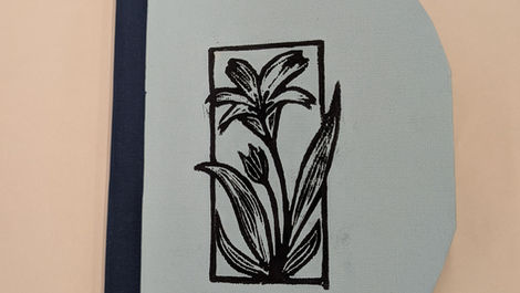 Small handmade book with a light blue, rounded cover, dark spine, and a centered black linocut flower design, with a green tab slightly visible from inside.