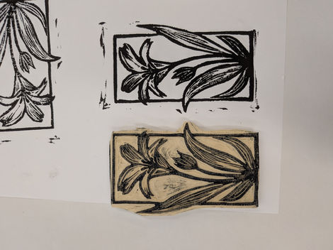 A hand-carved linoleum block rests on paper next to several black ink test prints of a floral design. The artwork features a stylized lily with long, flowing leaves enclosed in a rectangular border.