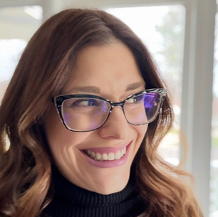 Our CEO Mandy is in a selfie, smiling looking off to the left. She is wearing a pair of black glasses and a black turtleneck. Her hair is medium brown and curled, and she is wearing a blue toned ink lipstick.