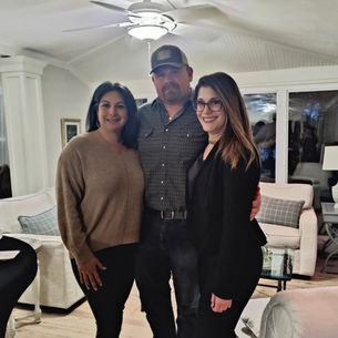 Mandy Krickhahn the founder of The Collective is standing oh the right side of a group photo with Steve Howe (center) and Jenn Howe (left) owners of Howe farms. They are standing in the center of an open living room with white walls and creme furniture. Jenn Howe is in leggings and a tan sweater, her hair is black and worn down in a middle part. Steve is wearing a blue button down with flannel print and jeans. He is also wearing a blue baseball cap and has facial hair directly around his mouth. Mandy is wearing a black cardigan and jeans, and has long brown hair parted on the side as well as thick framed black glasses. They are all smiling.