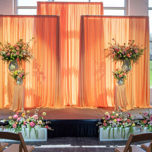 A picture of the altar SRE designed for the wedding featured in the magazine. It has three large panels of peach fabric, draped from rods so that the outside panels are slightly lower and cover the center panel of fabric. There are also two columns about waist height draped in the same peach fabric. They have two white flower vases with pink, yellow, peach, and orange flowers in it, with the highlight flower being peach peonies. surrounding the altar are white floral boxes with the same flowers inside and added greenery pieces that spill over the edge of the boxes. There are brown chairs on both sides of the aisle.
