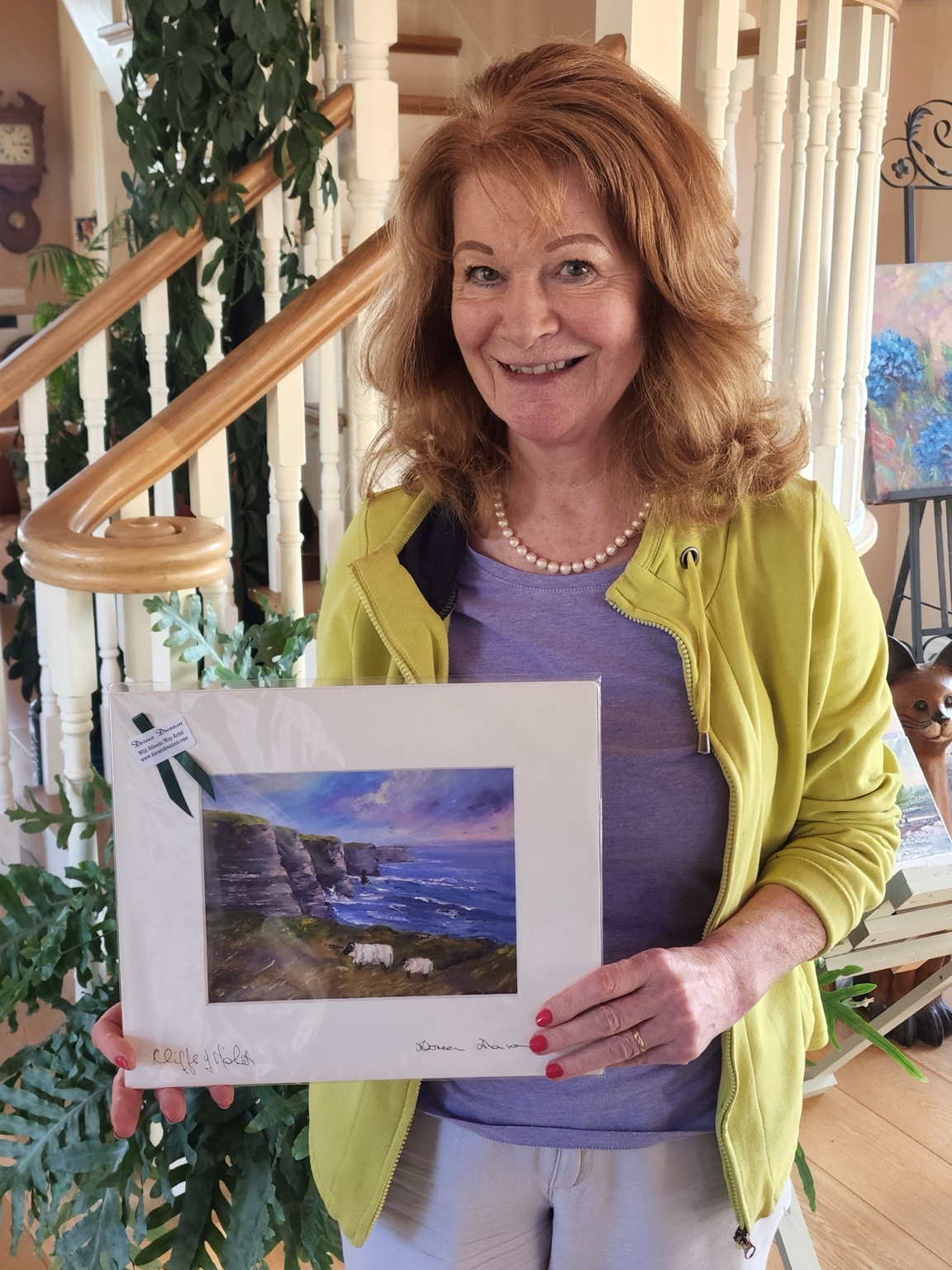 Irish artist Doreen Drennan smiling in her Doolin studio while holding her Cliffs of Moher coastal artwork beside a curved staircase.