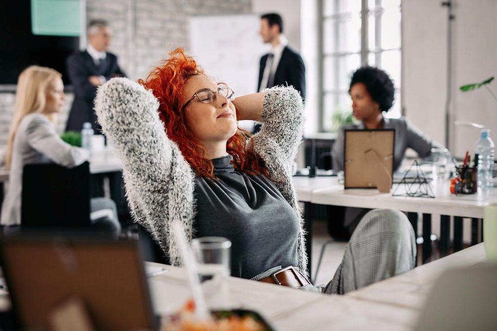 Woman with red hair relaxes at an office desk with her hands behind her head. Colleagues work in the background. Bright, casual setting.