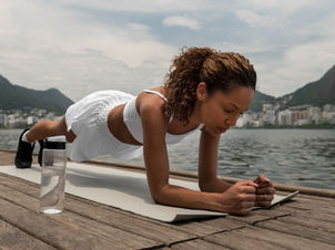 Woman in workout attire planking on a pier by a lake with city and mountains in background. A water bottle is beside her. Calm, focused mood.