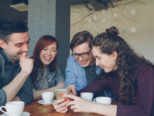 Four friends laughing and looking at a phone in a cozy café. They are surrounded by coffee cups, with warm lighting in the background.