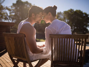 Couple seated outdoors on wooden chairs, foreheads touching, holding hands. Sunlit setting with trees in the background. Calm and intimate mood.