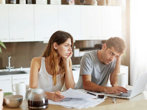 A woman and man look stressed while checking bills and using a laptop in a bright kitchen. Papers, a coffee pot, and mugs are on the table.