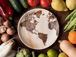 Wooden world map plate surrounded by fresh vegetables and fruits like tomatoes, lemons, and cucumbers on a dark wooden table.