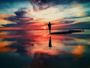 Silhouette of person with arms outstretched at sunset, standing on a reflective beach. Vibrant orange, pink, and blue sky. Tranquil mood.