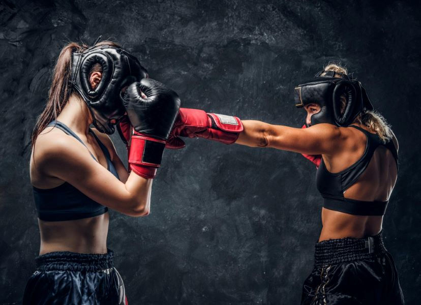 Two women in boxing gear spar in a dark gym. One lands a punch, both wear black headgear and gloves, creating a tense atmosphere.