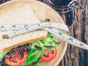 A sandwich with lettuce and tomato on a wooden plate, wrapped with a measuring tape, suggesting a focus on diet or health. Rustic table.