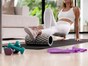 Woman in white activewear using a foam roller on a yoga mat in the living room. Pink shoes, teal dumbbells, and purple resistance band visible. Bright setting.