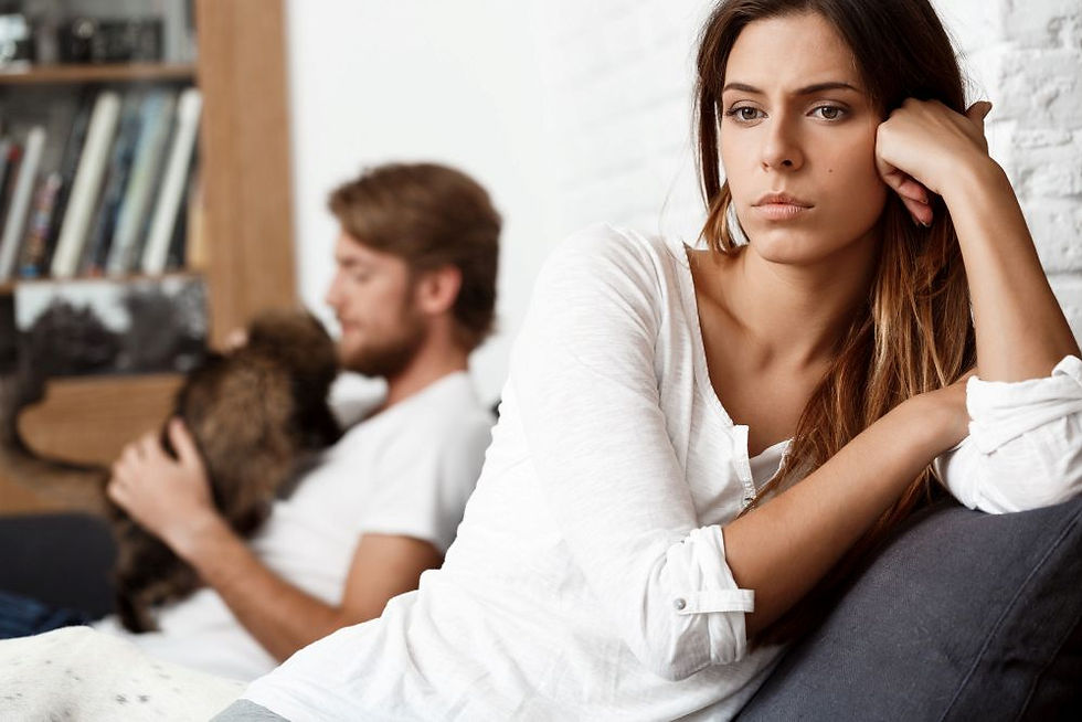 Woman in white shirt looks thoughtful, leaning on arm, while man in background holds a dog. Shelves with books are visible. Calm mood.