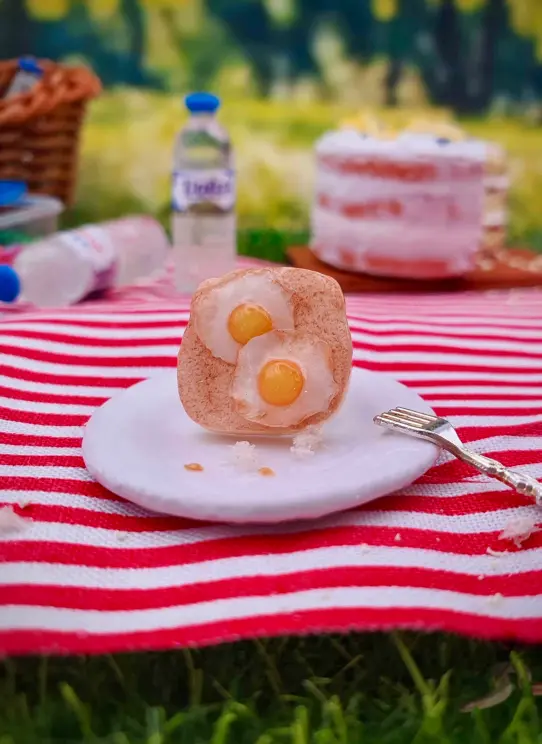 Miniature clay eggs on toast pin and various food on a red and white picnic blanket
