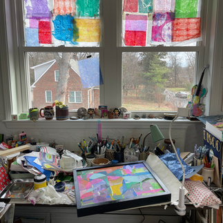 Molly's table in the studio bathed in beautiful light from the window. Colorful papers cover the top half of the window and lots of art supplies and one framed abstract piece are on the desk.
