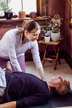 Female yoga teacher assisting a male yoga student lying on the ground in an assisted bridge position. 