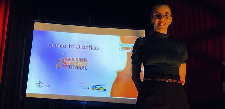 Foto da intérprete de Libras Vanessa Fernandes Nascimento com Telão com a logo marca do PIC e título Concerto Didático e logomarcas do Ministério da cultura, Governo Federal