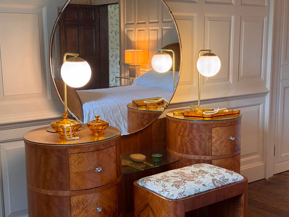 The dressing table in  Mrs Harmsworth's bedroom at Athelhampton House. It's in art deco style, with a large circular mirror framed by two circular sets of drawers. In between the drawers is a small side with a matching jade-handled mirror, hair brush, and hair box set. On each of the sets of drawers is a large, spherical light, and some plastic boxes reminiscent of the shape of sea shells in a golden brown colour, containing strings of pearls. A stool upholstered in cream and floral brocade sits in front of the dressing table. The mirror reflects an art deco bed with white sheets, and a door opening into another room. The walls are wood panelling painted cream.