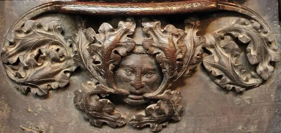 A very round, wooden carved face with large oak leaves issuing from the wide open mouth to frame the face both top and bottom as well as swirling around either side of the face.