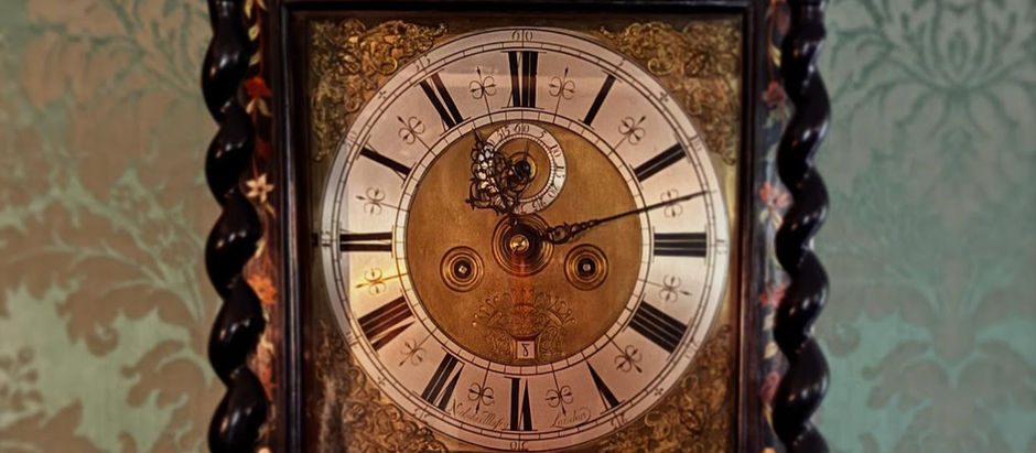 A grandfather clock face against green silk damask wall coverings. The frame of the clock is dark wood, inlaid with flowers and leaves in pale green, white, and dark orange. The face of the clock is mostly gold, with the numbers in roman numerals against silver. The time shown is 11:13.