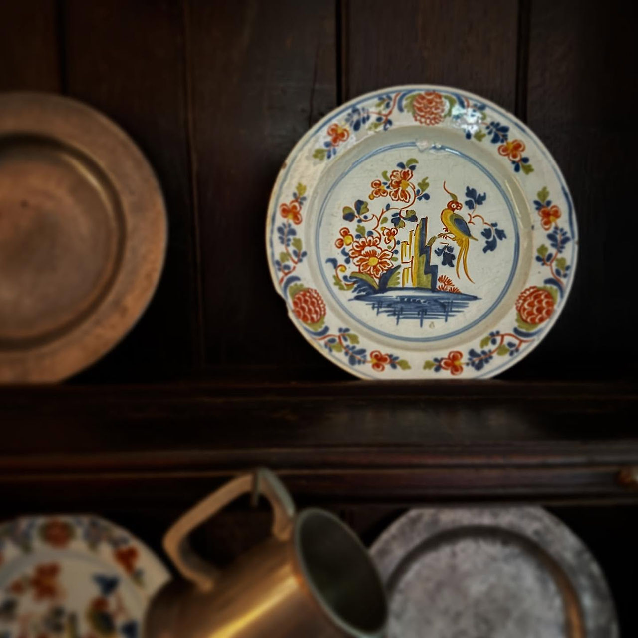 A painted 18th century plate, decorated with flowers around the rim and thn flowers and a bird in the middle. The glazes are red, green, yellow, and blue. It sits on a dark wood dresser surrounded by other matching plates and pewter plates.