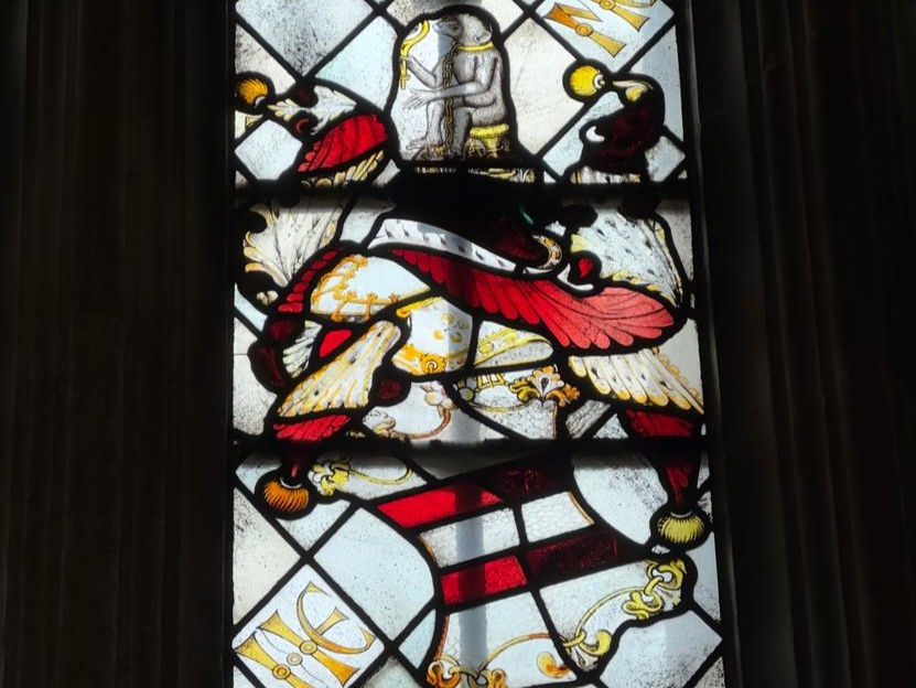 Stained glass window showing a shield with five horizontal stripes alternating white and red. It's tilted at an angle, and has a helmet perched on top. On top of the helmet sits an ape gazing into a hand mirror. The helmet has feathers spiralling out of it in red and white