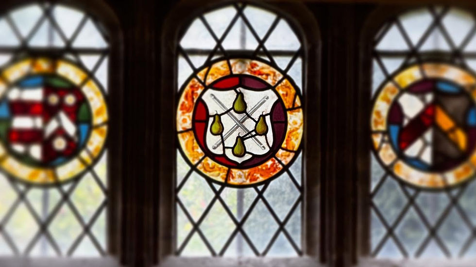 Glass in the Great Chamber at Athelhampton. The central roundel is in focus, showing a white shield with four pears and crossed grozing irons dividing them - the Kelway coat of arms. The background of the roundel is red, and it's surrounded by orange and white decoration as a border.