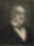 A black and white photograph of a severe, slightly sad looking man with thinning hair and mutton chops. He's on the older side, with a heavily lined face. He has a high, stiff, white collar and a bow tie with small white polka dots. He has a white waistcoat under a dark coloured coat.