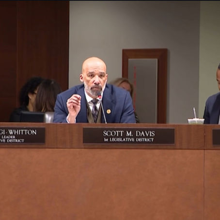 Scott M. Davis, Legislator for the 1st District, speaks into a microphone during a Nassau County Legislative meeting. Seated beside him are Delia DeRiggi-Whitton, Minority Leader of the 11th District, and Carrié Solages, Legislator for the 3rd District. They appear focused and engaged in session.