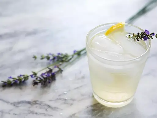 A glass with ice, lemon peel, and a lavender sprig on a marble surface, creating a refreshing and calm setting. Lavender flowers nearby.
