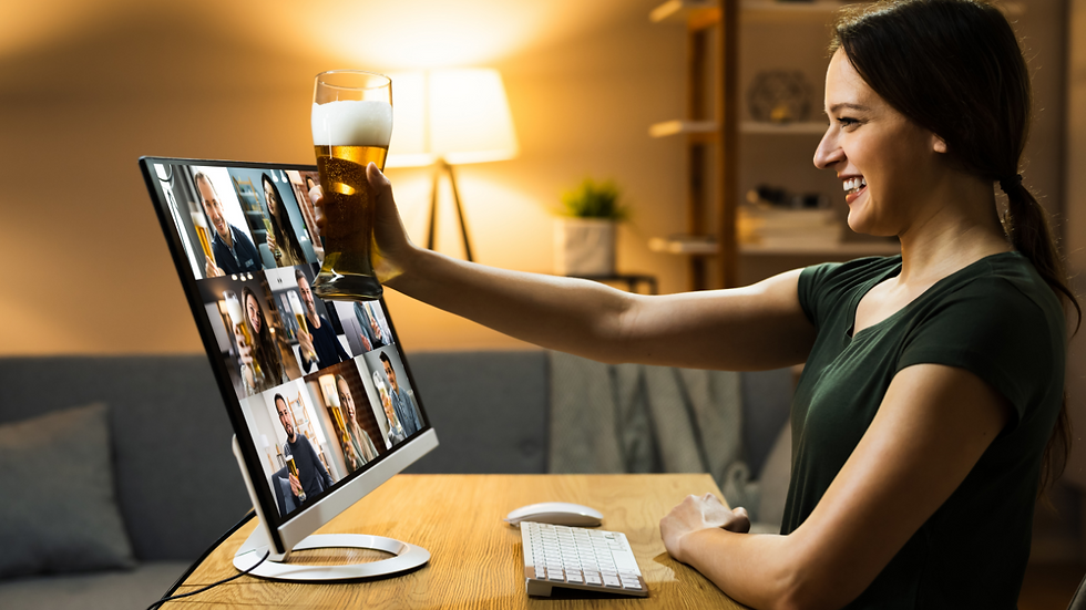 Woman in a dark green shirt toasts with beer during a virtual meeting on a monitor. Warm lamp light, cozy room, smiling, happy mood.