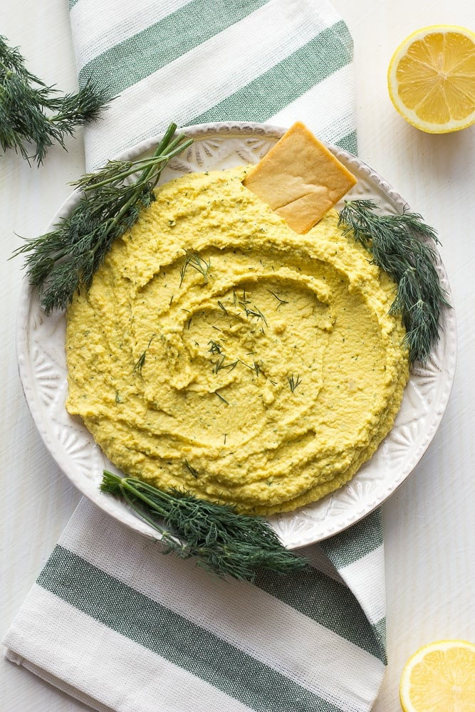 A plate of creamy yellow hummus with dill and a cracker on top. Set on a green-striped cloth, with lemons and dill sprigs beside.