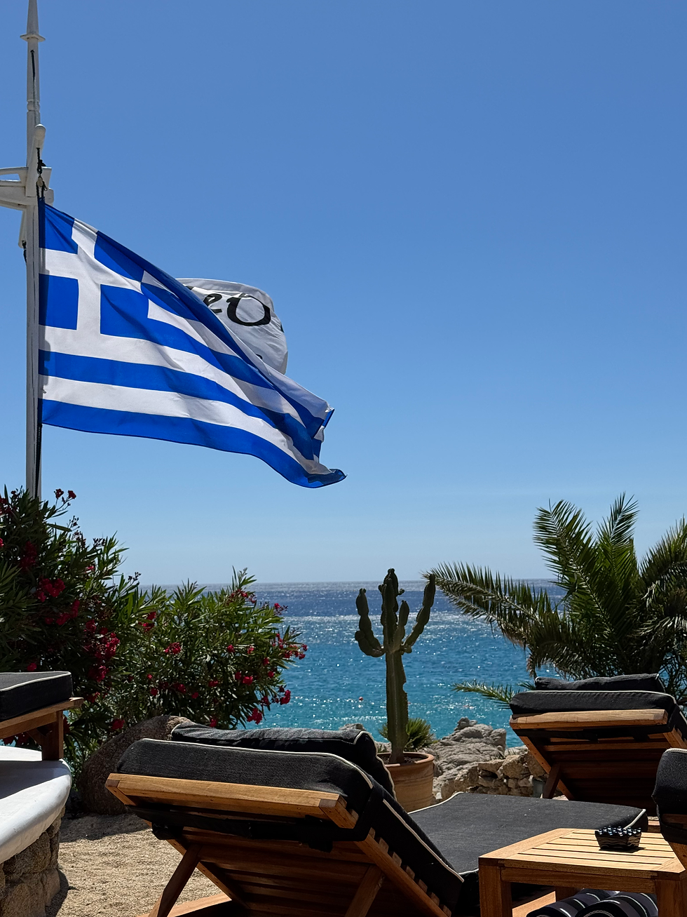 Greek flag waving with a view of the sea, sun loungers, and tropical plants in the foreground.