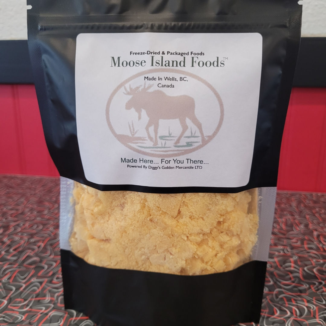 "Black package of freeze-dried food from Moose Island Foods, showcasing its contents through a clear window, which appear yellowish and crumbly. The label reads: "Freeze-Dried & Packaged Foods Moose Island Foods™ Made in Wells, BC, Canada Made Here... For You There... Includes production credits: 'Powered by Diggy's Golden Mercantile LTD.'"