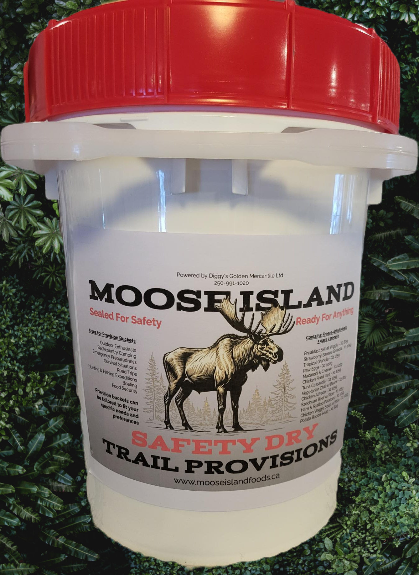 A white plastic container with a red lid. The label reads 'MOOSE ISLAND SAFETY DRY TRAIL PROVISIONS,' featuring a moose illustration. Additional text includes 'Sealed For Safety' and 'Ready For Anything,' along with 'Powered by Diggy's Golden Mercantile Ltd.' A website URL (www.mooseislandfoods.ca) is also included. The background showcases green foliage