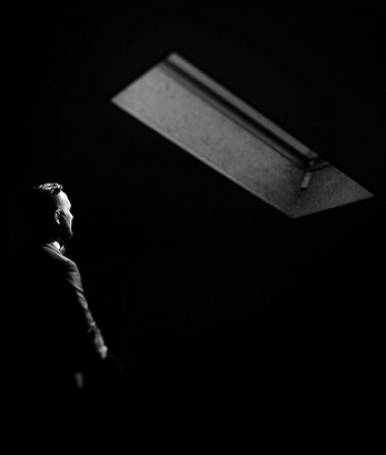 A businessman in a suit stands in a dark room, looking up at a single skylight of light.