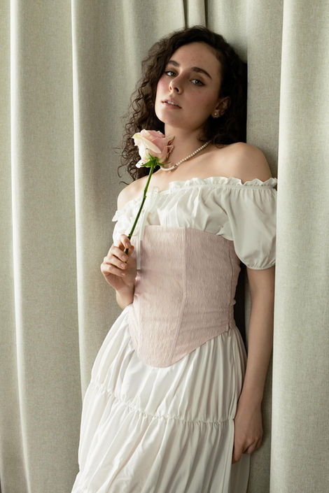 ditorial fashion portrait of a model in a soft, vintage-inspired white dress and corset, holding a single rose against a minimalist curtain backdrop
