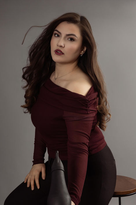 Professional branding portrait by a fashion photographer shows a female hair stylist in a maroon top holding a blow dryer as her own long, wavy hair blows. Dramatic studio side lighting highlights the action on a neutral grey backdrop, capturing motion and style
