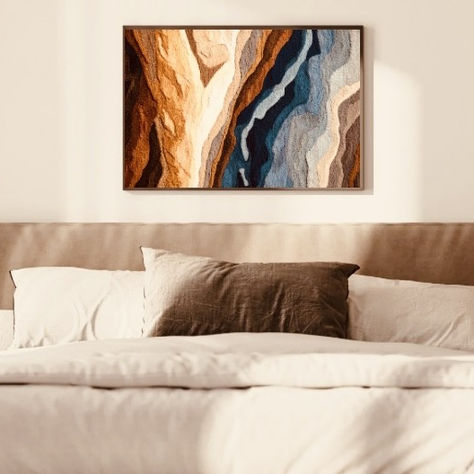 Textured fiber art with flowing geological layers in deep blues, sandy creams, and warm browns, framed in dark wood and styled above a soft, neutral-toned bed.