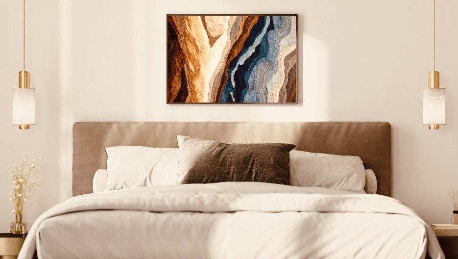 Textured fiber art with flowing geological layers in deep blues, sandy creams, and warm browns, framed in dark wood and styled above a soft, neutral-toned bed.